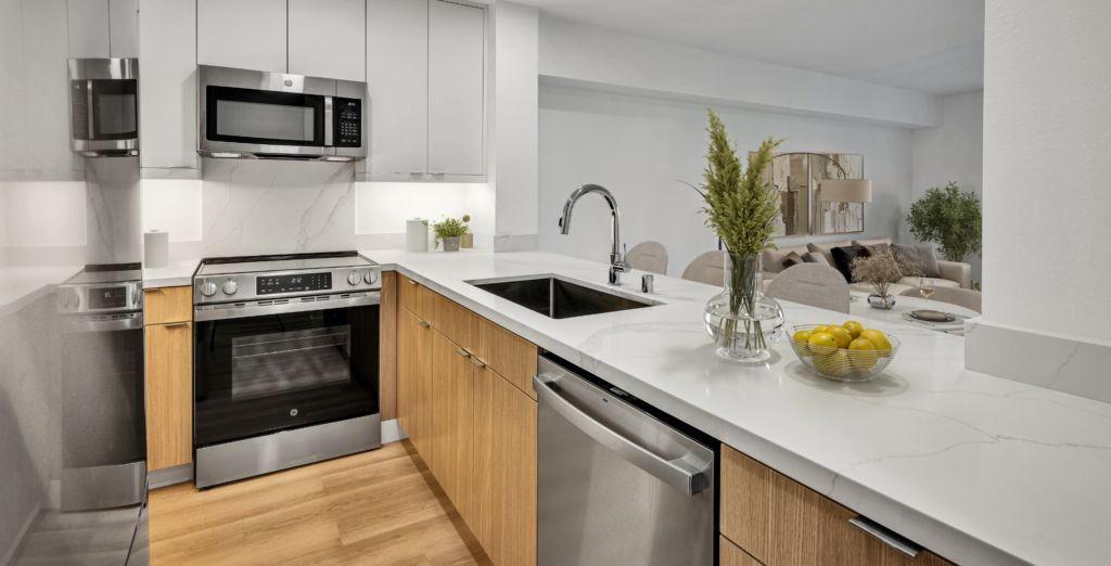 Open-concept kitchen with white countertops, wood cabinetry, stainless steel appliances, and living area beyond.