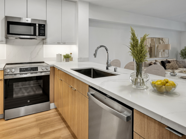 Open-concept kitchen with white countertops, wood cabinetry, stainless steel appliances, and living area beyond.