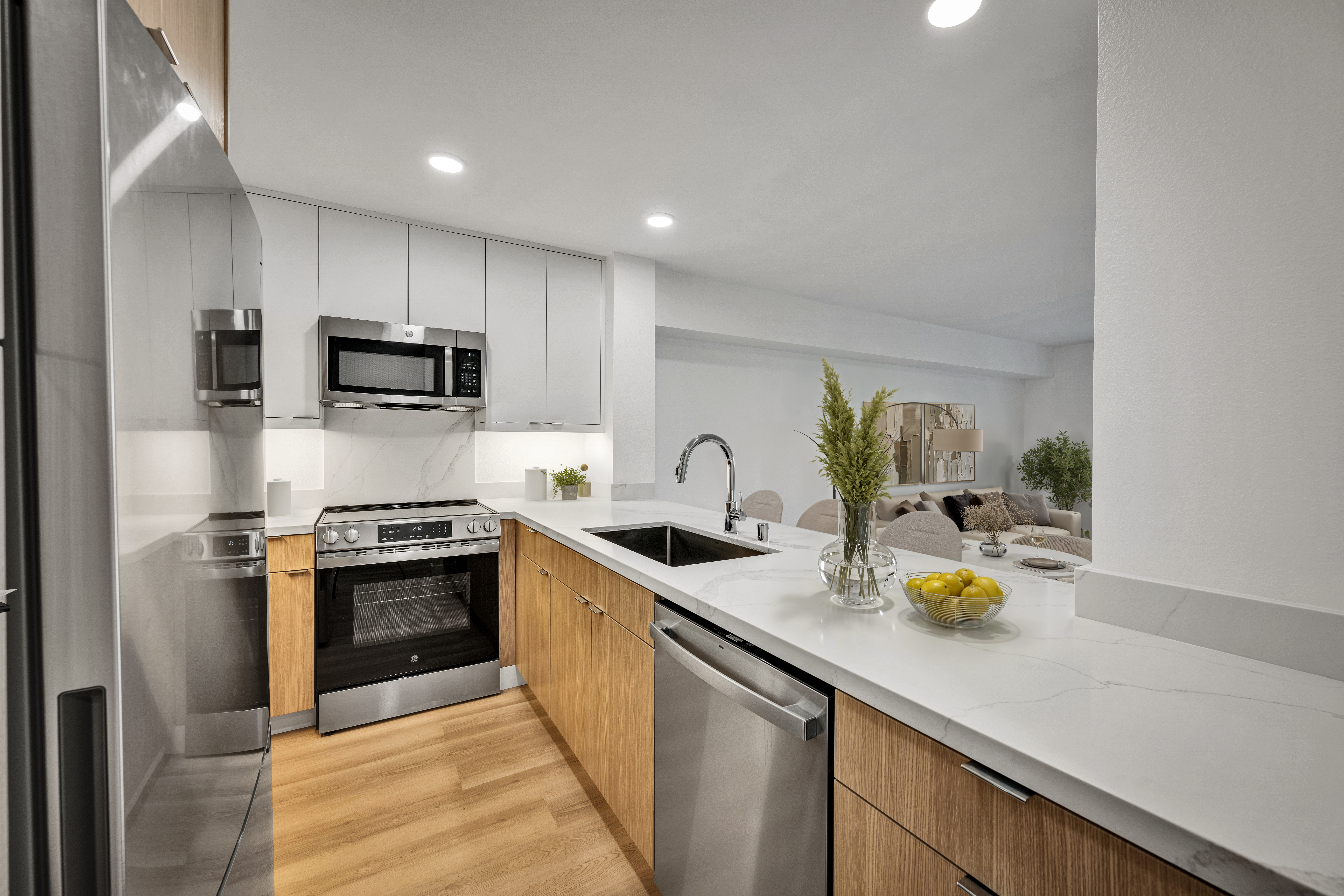 Open-concept kitchen with white countertops, wood cabinetry, stainless steel appliances, and living area beyond.
